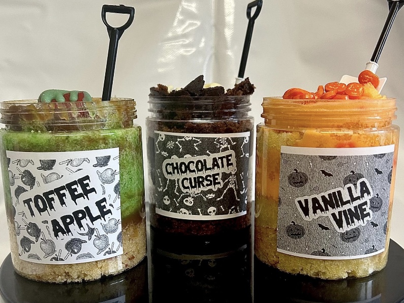 Halloween cake jars labelled Toffee Apple, Chocolate Curse and Vanilla Vine on a black stand