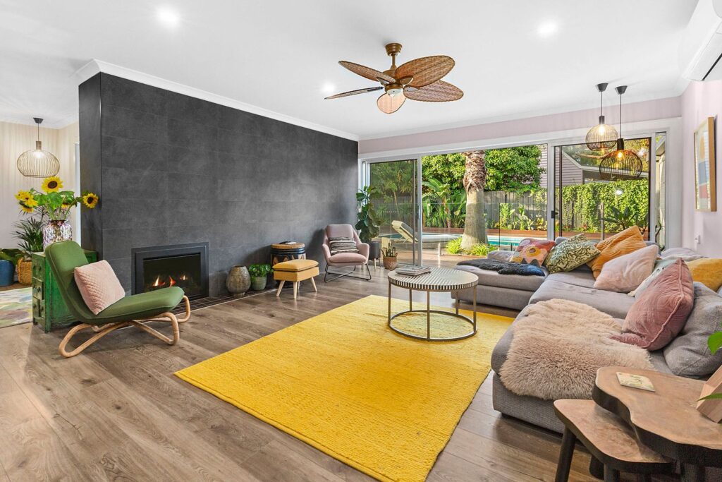 Eclectic living room in Australia — mustard yellow rug, green rattan chaise, black feature wall and rattan pendant lights with garden views