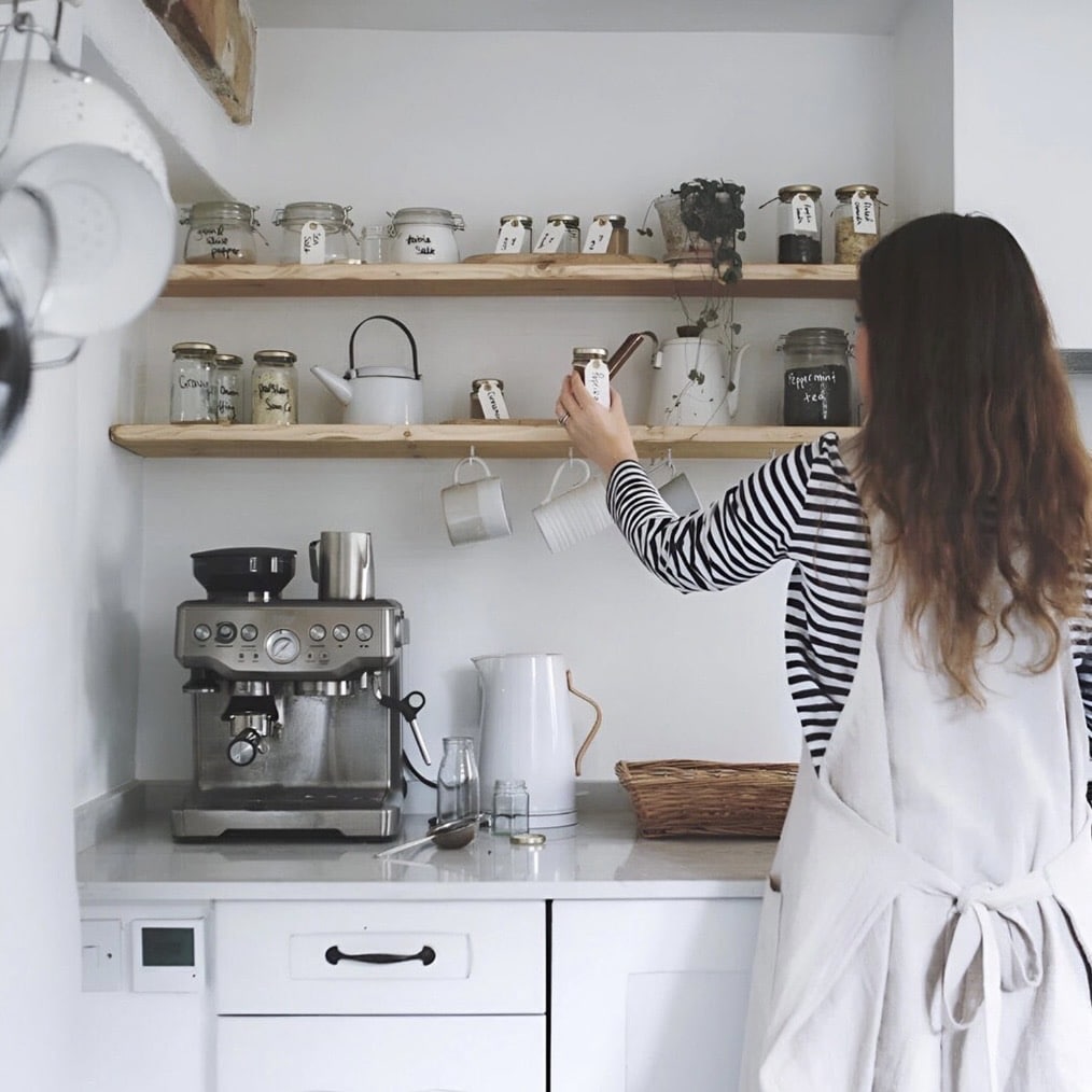 Rustic wooden kitchen floating shelves above an espresso machine, holding labelled glass storage jars, tea tins and a white enamel kettle, with mugs hanging on hooks beneath