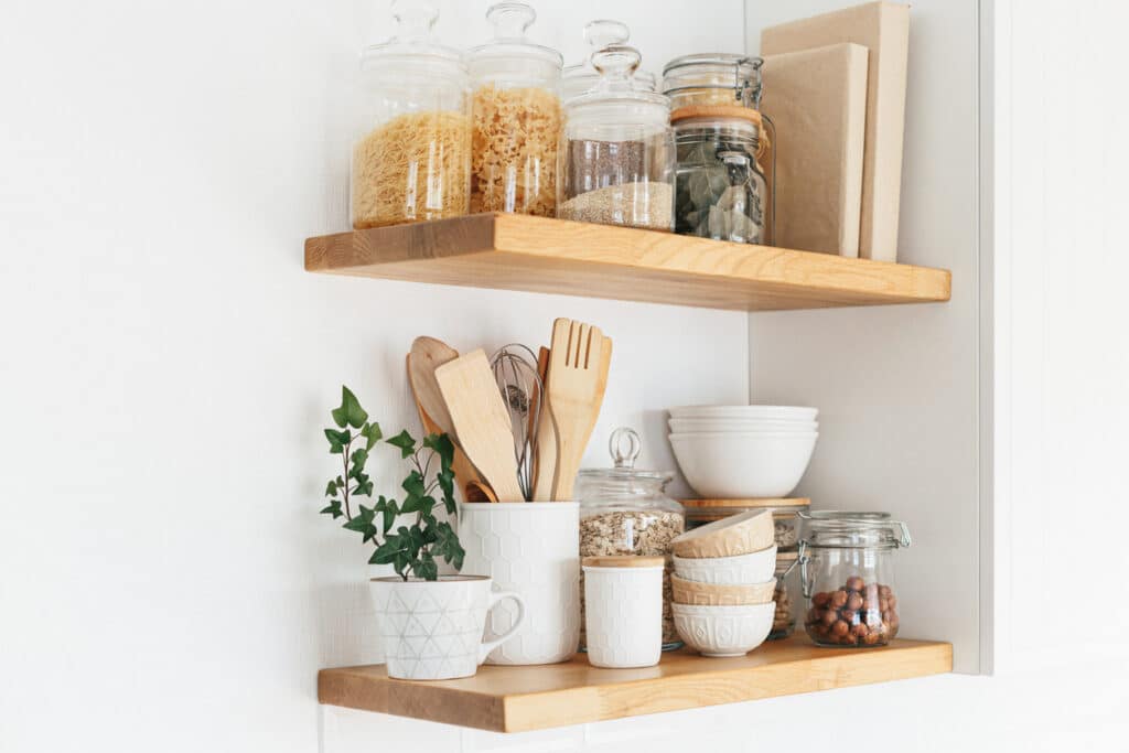 Two wooden floating shelves holding glass storage jars of pasta and grains, white ceramic bowls, wooden utensils and a trailing ivy plant