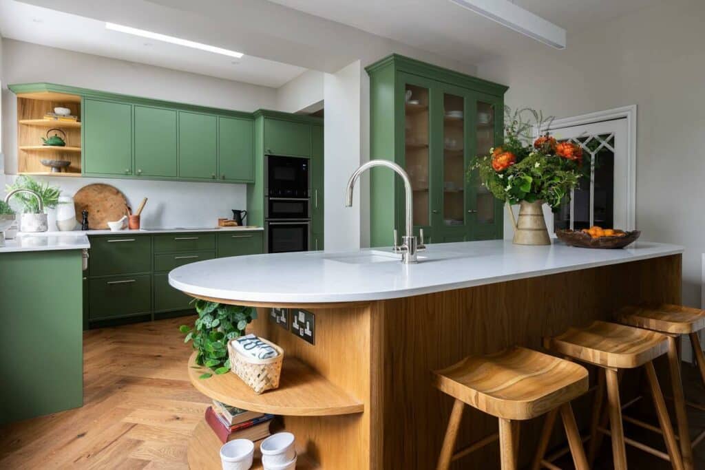 Two tone kitchen with Farrow & Ball Calke Green perimeter cabinets, curved natural oak island with saddle bar stools and herringbone oak flooring.