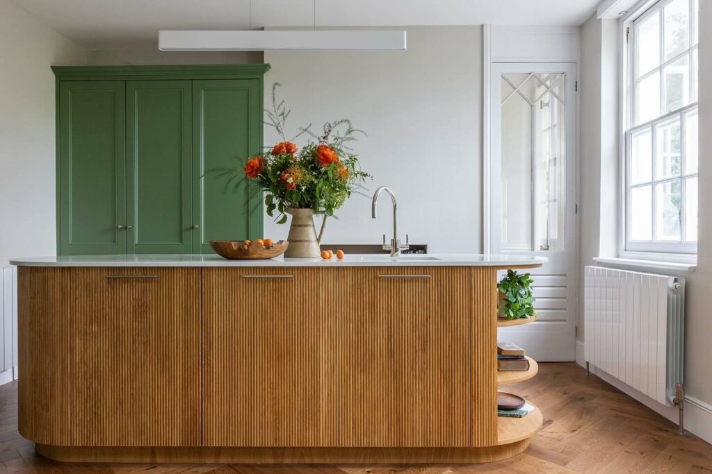 Curved reeded natural oak kitchen island with white marble worktop and Farrow & Ball Calke Green pantry cupboard behind.​​​​​​​​​​​​​​​​