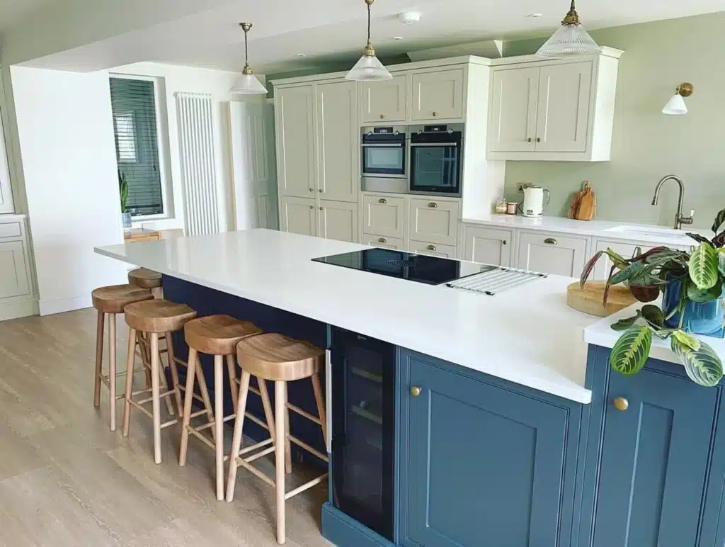 Two tone kitchen with cream shaker perimeter cabinets, deep navy island, pale grey-green walls and natural beech saddle bar stools at the breakfast bar.​​​​​​​​​​​​​​​​
