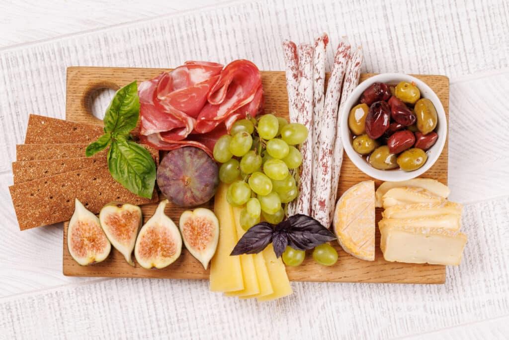 Cheese platter accompaniments including prosciutto, figs, grapes, olives and crackers