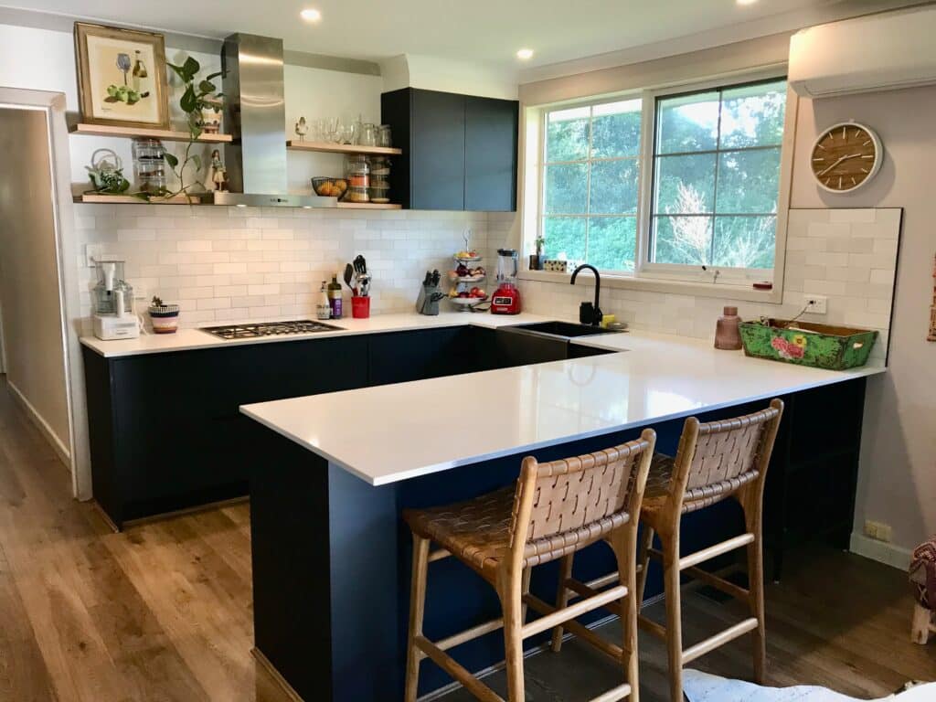 Kitchen renovation after with handleless navy units, a white quartz peninsula, leather bar stools, open oak shelving and white brick-style tiles