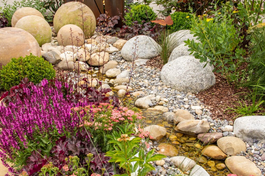 Low maintenance garden idea with water, pebbles, boulders, and a shallow rill surrounded by flowers.