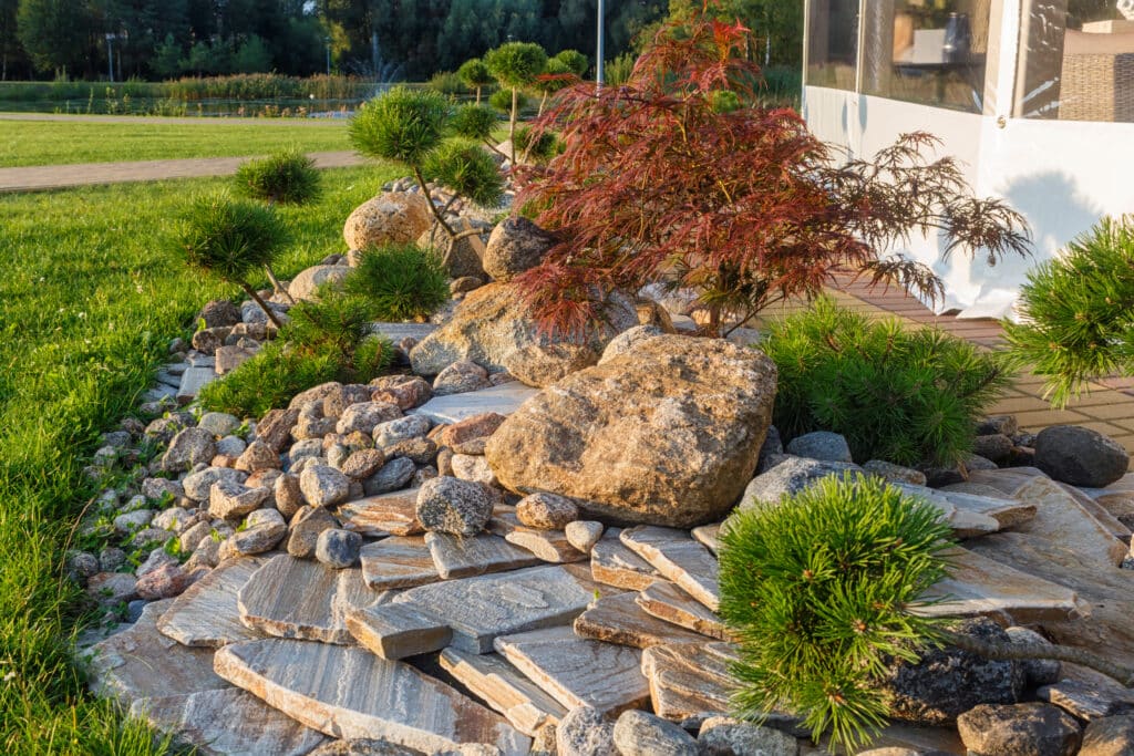 Low maintenance modern garden ideas: rock garden with boulders, layered stone paving and dwarf pines beside a patio.