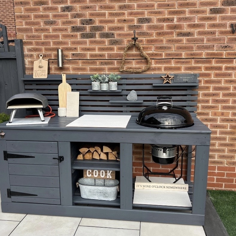 Grey painted wood outdoor kitchen with built-in Weber kettle barbecue, pizza oven and log storage against a brick wall