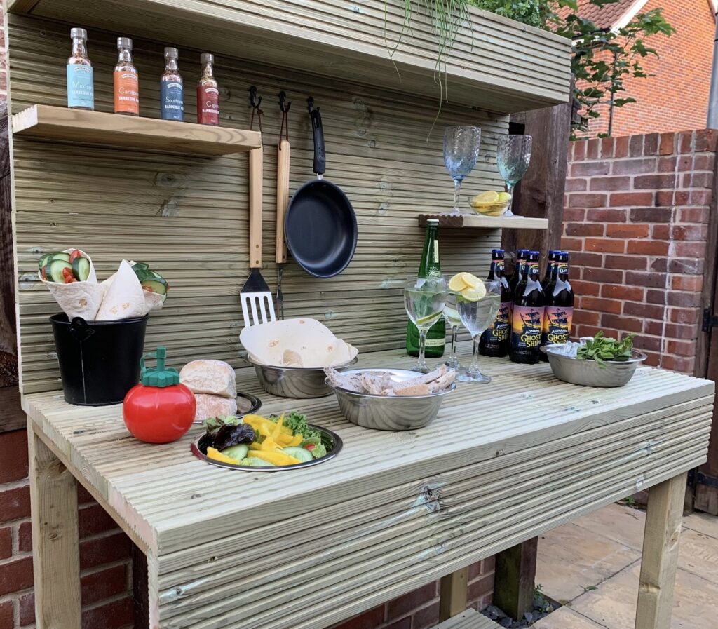 Natural wood outdoor kitchen bench with hanging utensil hooks, upper shelving and worktop styled for al fresco dining
