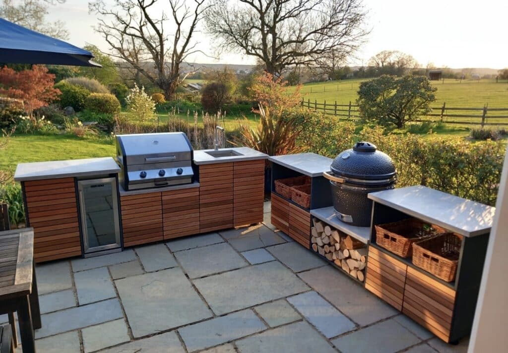 Modular outdoor kitchen with timber-clad cabinetry, built-in gas grill, ceramic egg-style barbecue and log storage, overlooking open countryside