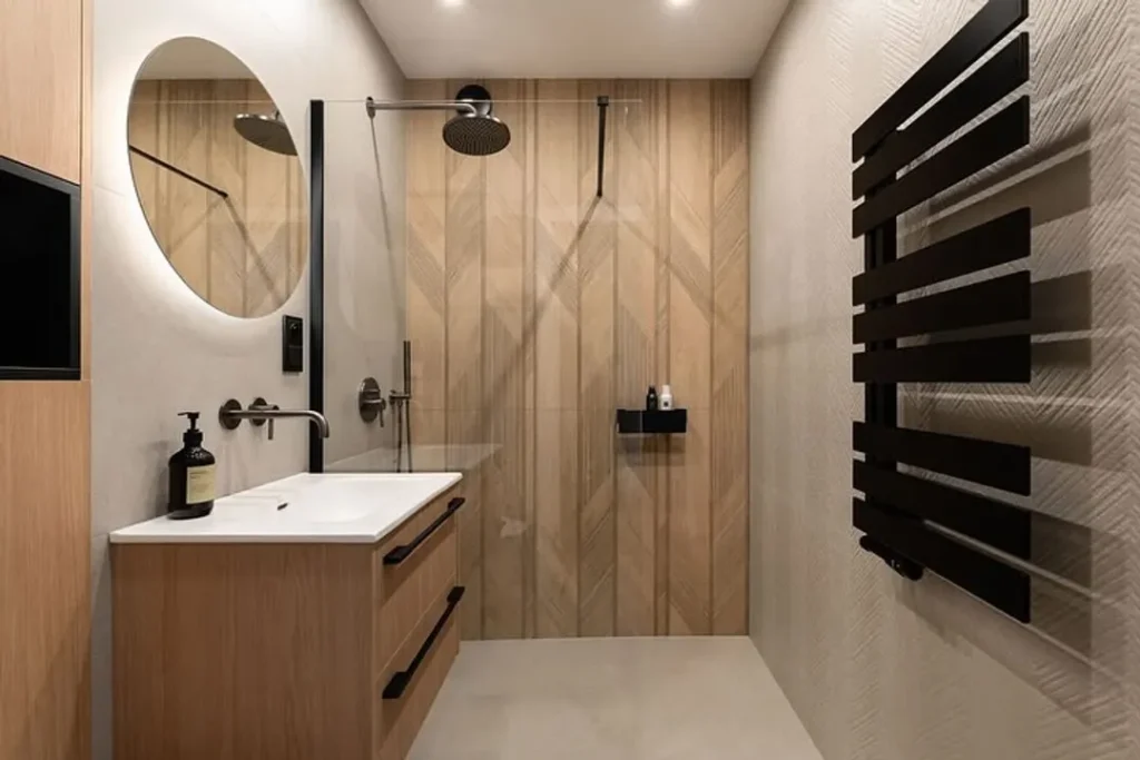 Small walk-in shower with chevron wood-look tile, round mirror, black fixtures and black towel radiator