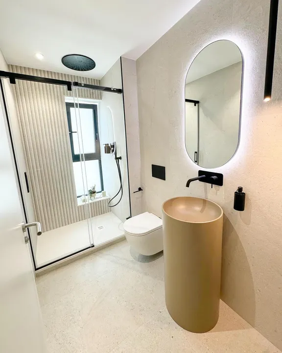 Bathroom with sliding barn-door shower screen, sand-tone pedestal sink, backlit oval mirror and Porcelanosa Durango Bone ribbed tiling