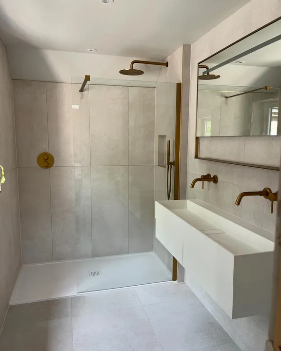 Walk-in wet room shower with large-format grey tiles, brass fittings and a floating trough sink
