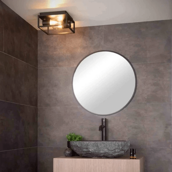 Cage-style flush ceiling light above a round mirror and stone vessel sink in a dark tiled bathroom