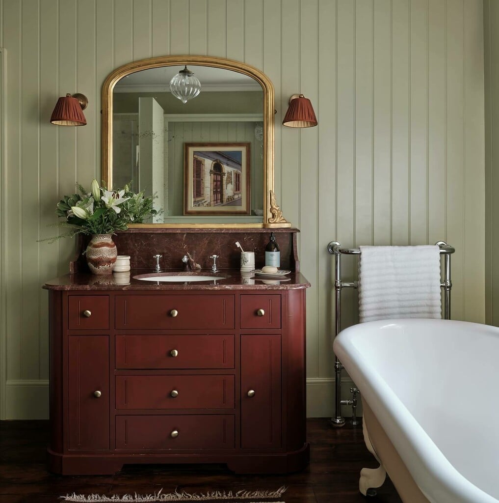 Pleated fabric wall sconces either side of a gilt mirror above a burgundy vanity in a sage green panelled bathroom