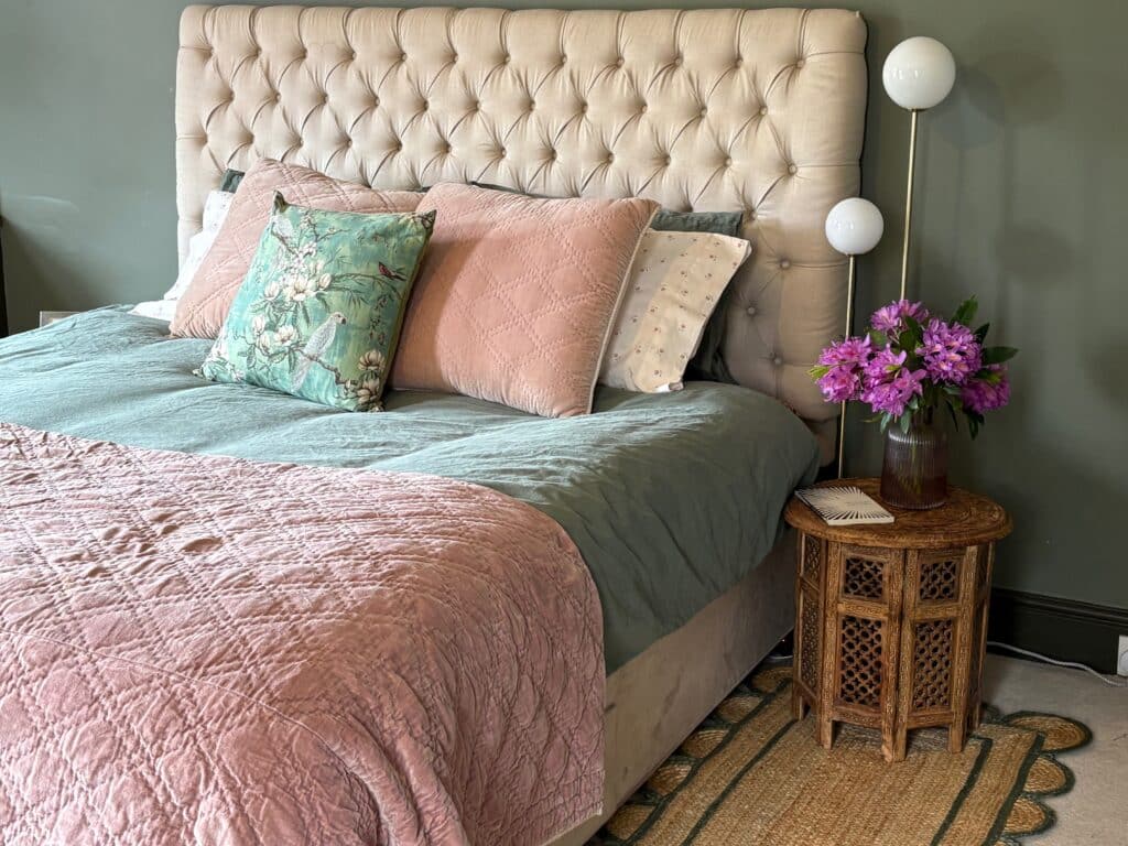 Pink and green bedroom for adults with a blush velvet quilt over sage green bedding, a chinoiserie parrot cushion, a carved wooden side table and a jute rug with a green scalloped border