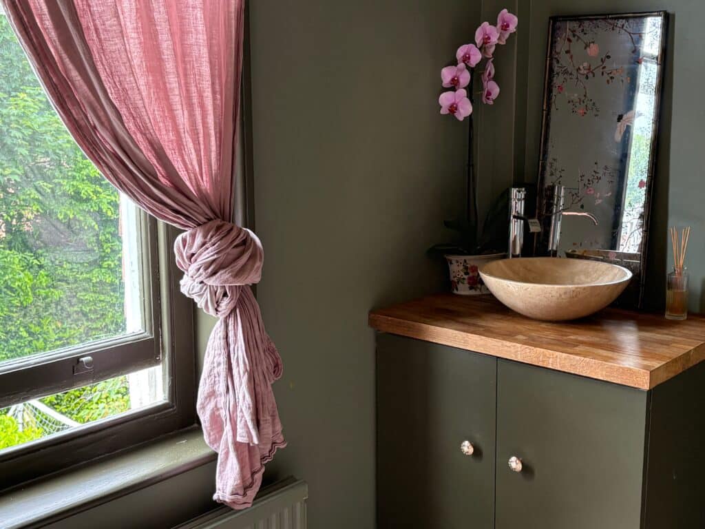 Green and pink bedroom washbasin corner with a stone vessel basin on an oak worktop, a knotted pink linen curtain and a chinoiserie mirror panel
