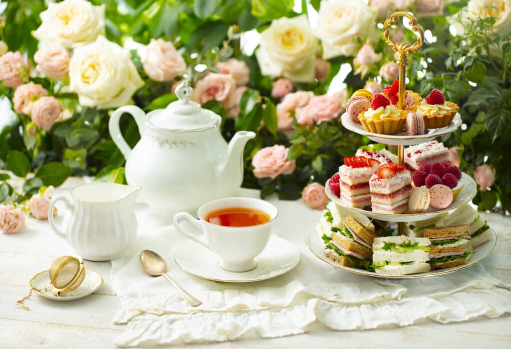 Three tier stand with finger sandwiches, layered cakes, fruit tartlets and macarons beside a white teapot and cup of tea