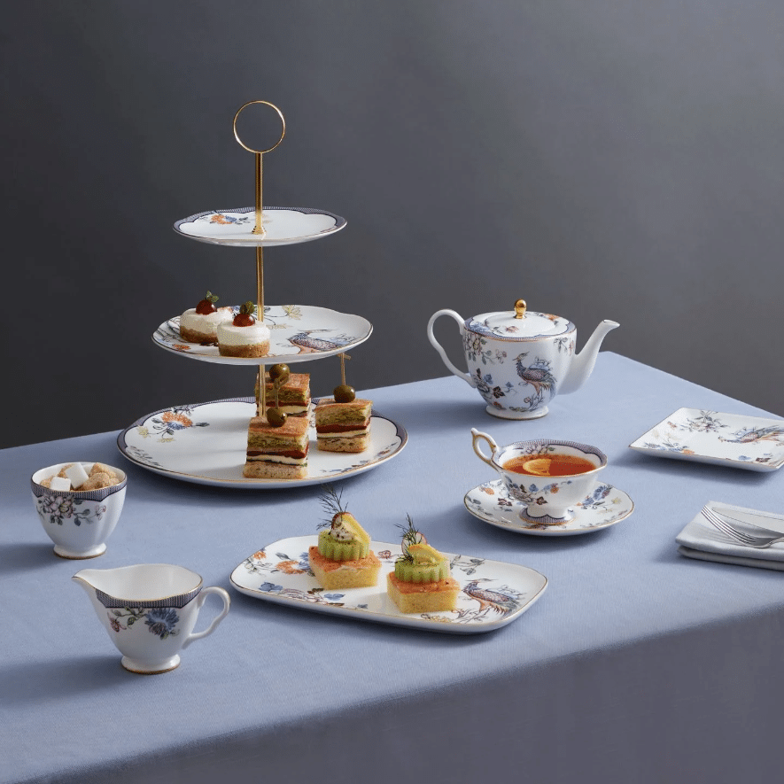 Afternoon tea table with floral Wedgwood teapots, a tiered stand of scones and finger sandwiches, and a cup of tea on a blue-grey cloth