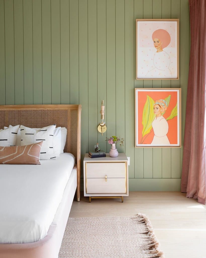 Sage green panelled bedroom wall painted in Farrow & Ball Lichen with pink and coral art prints, a brass wall light and a dusky pink velvet curtain