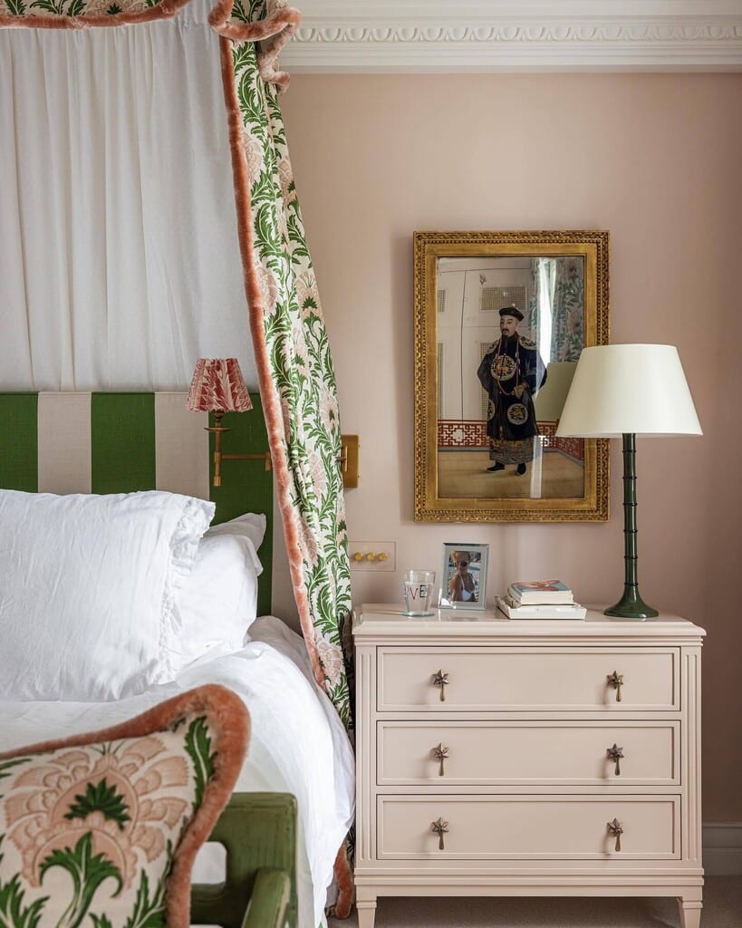 Pink and green bedroom with Farrow & Ball Pink Ground walls and bedside chest, a green and white striped headboard, chintz bed curtains and a green lamp