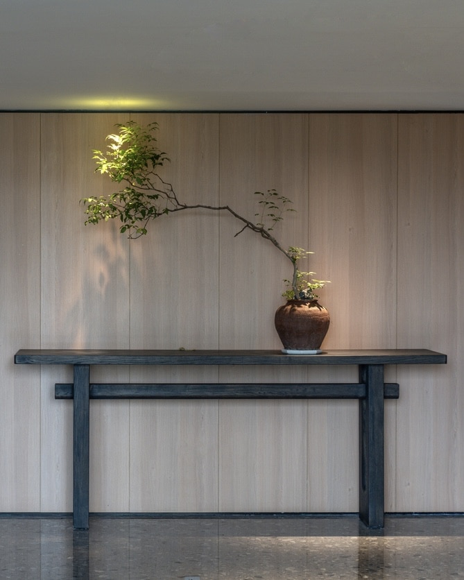 Black trestle-style console table against pale wood wall panelling, styled with a single ceramic pot and dramatically branching tree, polished terrazzo floor