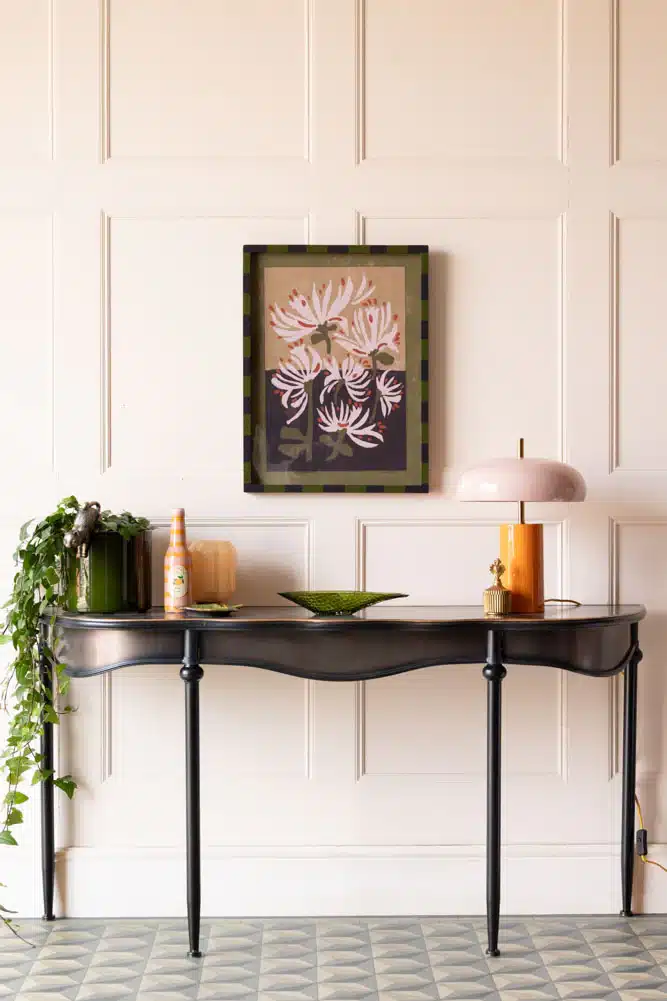 Black and gold antiqued console table with scalloped apron and tapered legs, styled with a dusky pink dome lamp, trailing ivy and amber glass vessels against a panelled wall