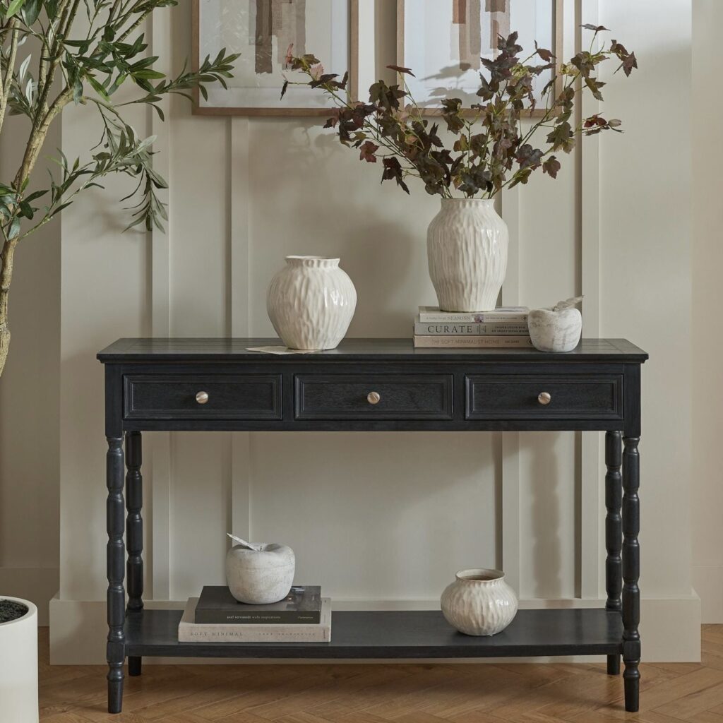 Black wood console table with three drawers and bobbin-turned legs styled with white ceramic vases, dried autumn branches and books on a lower shelf in a hallway