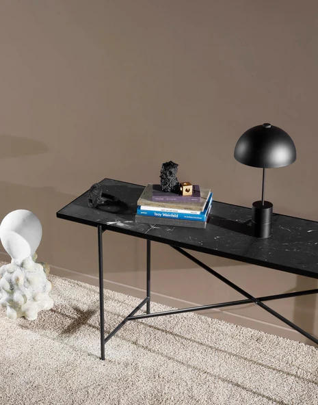 Black marble console table with slim black X-frame legs styled with a dome lamp, art books and a raw mineral sculpture against a warm taupe wall