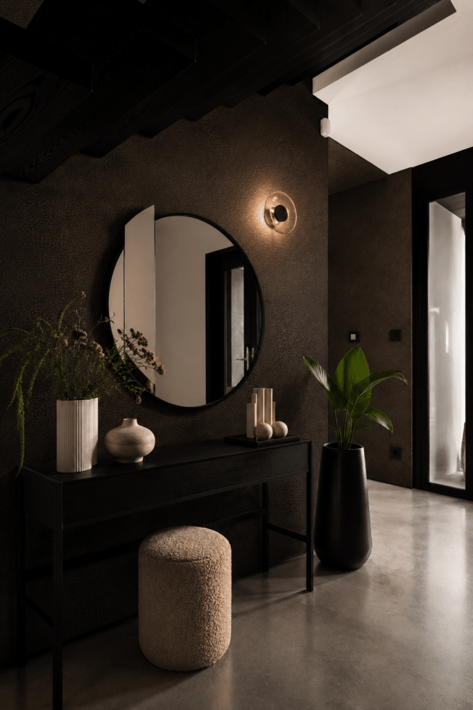 Dark hallway with black console table styled with a round mirror, ribbed ceramic vase, wild flowers and a large black floor planter against textured dark walls