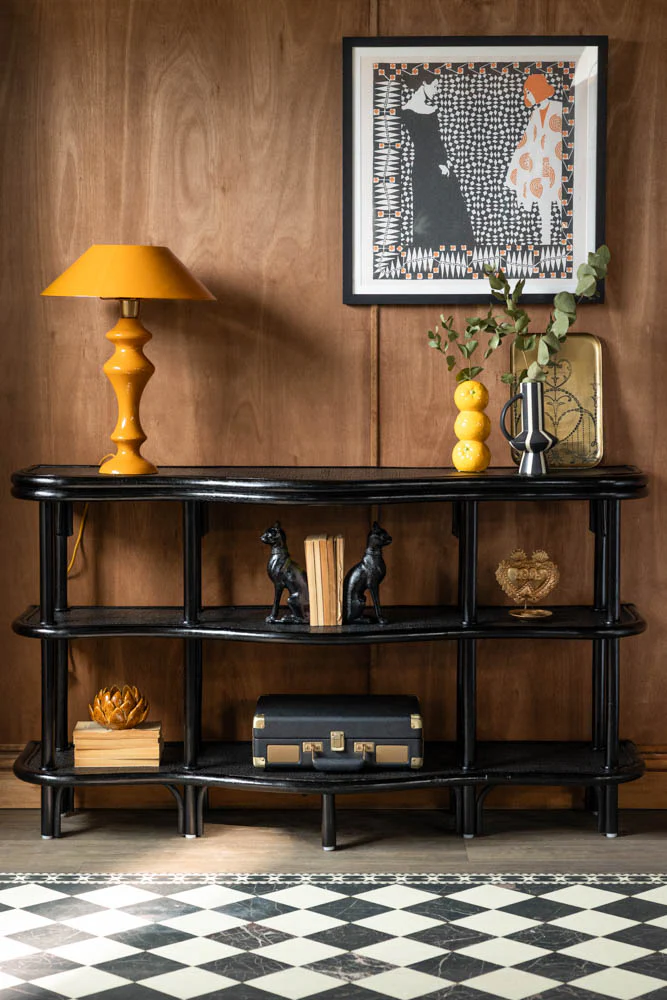Vintage-inspired black rattan console table with curved multi-tier frame, styled with an amber candlestick lamp, stacked ceramic vessels and a vintage suitcase on the lower tier, chequered tile floor