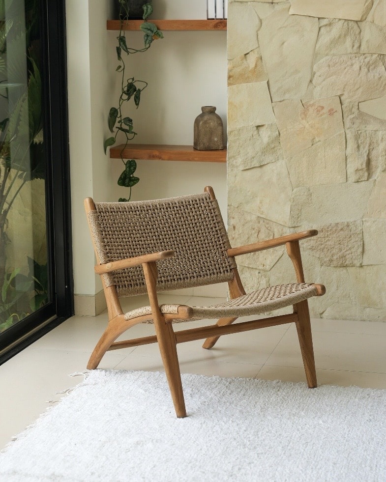 Handcrafted rattan accent chair with a teak frame beside a natural stone wall and trailing plant