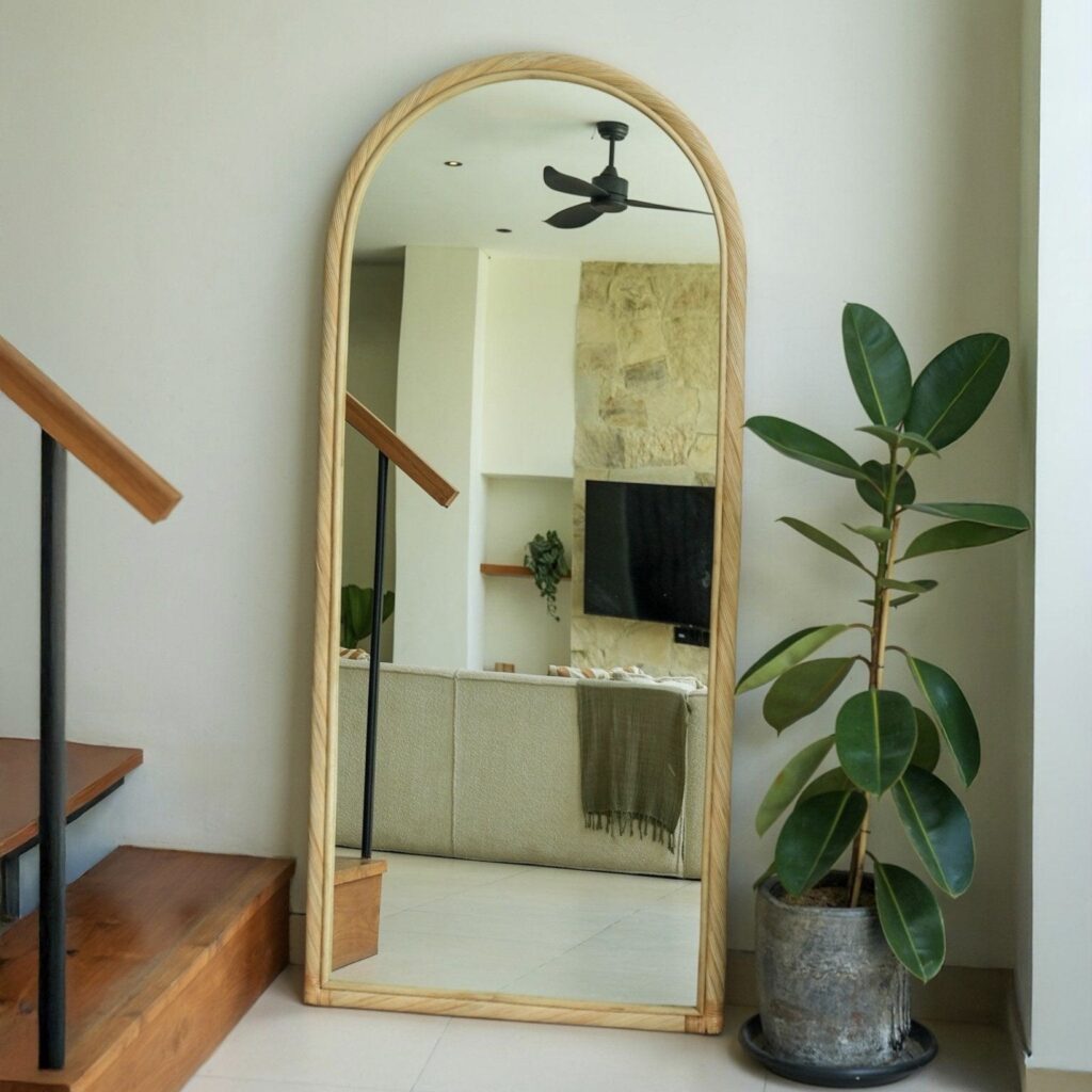 Wabi sabi mirror with an arched rattan frame leaning against a wall beside a rubber plant