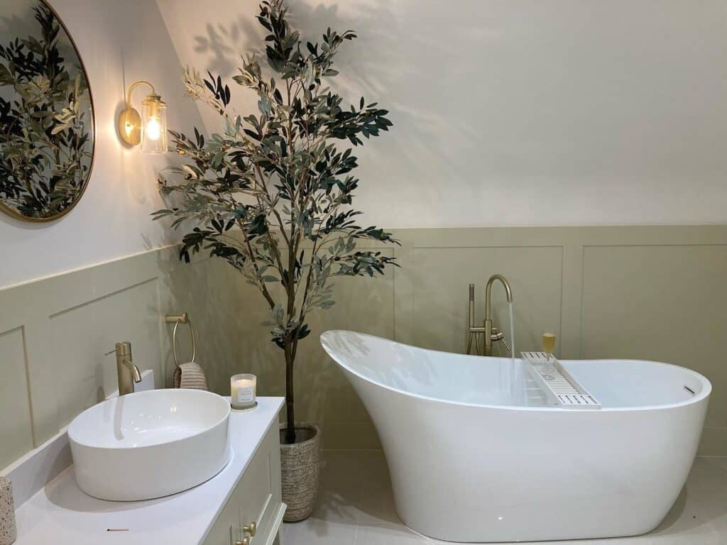 Bathroom with half-wall shaker panelling in Farrow & Ball Off-White, freestanding white bath, brass wall light and olive tree.