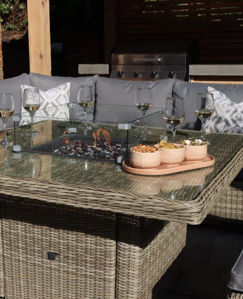 Close-up of glass wind guard on square fire-pit dining table with flame and glasses.