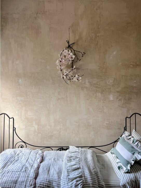 Green and beige bedroom with a wrought iron bed frame, striped bedding, ruffled accents, and a floral wall arrangement on a textured beige wall.