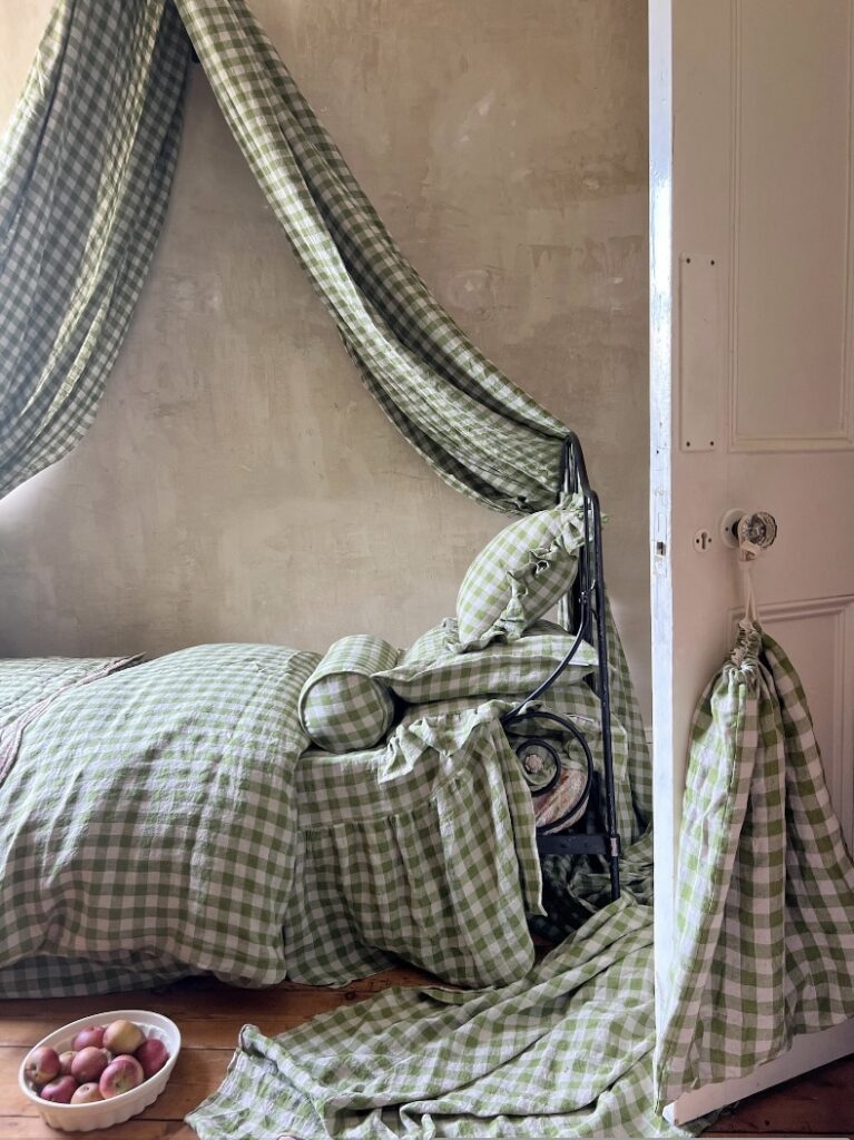 Green and beige bedroom with gingham bedding, draped fabric canopy, wrought iron bed frame, and a rustic bowl of apples on wooden flooring.