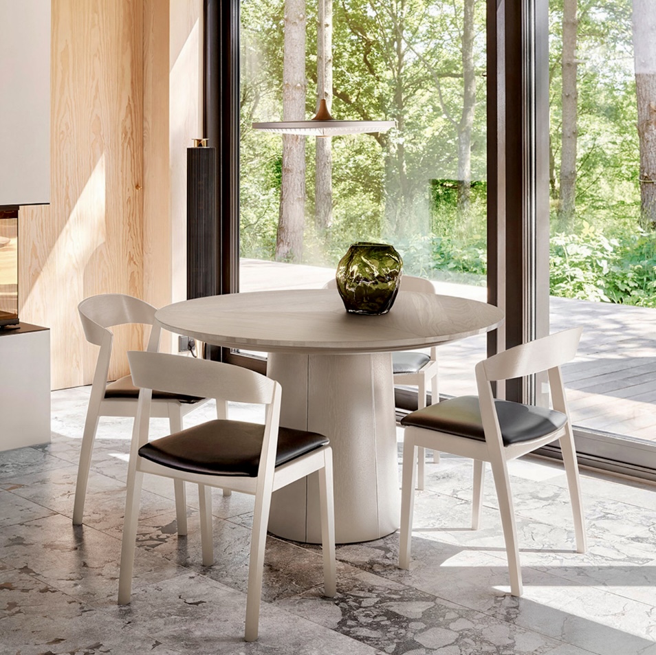Contemporary round extendable dining table in a pale oak finish, styled with minimalist white chairs and set against floor-to-ceiling windows in a serene, nature-inspired dining space.