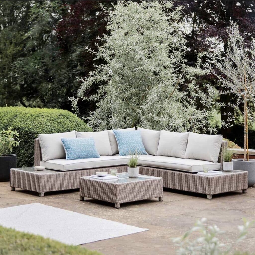 Woven rattan garden corner sofa with neutral cushions and square coffee table on a patio with greenery