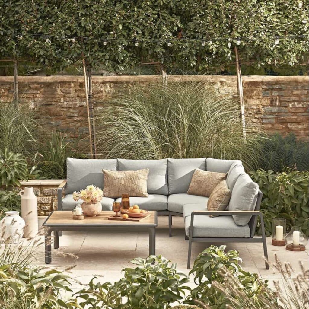 Corner garden sofa with durable aluminium frame, wood-effect detailing and neutral cushions on a patio with greenery and a stone wall
