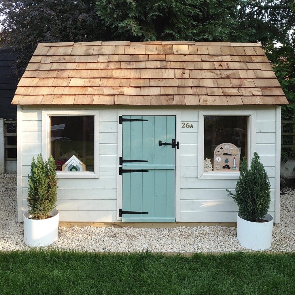 Cottage-style wooden playhouse with cedar shingle roof and pale blue door in a garden setting