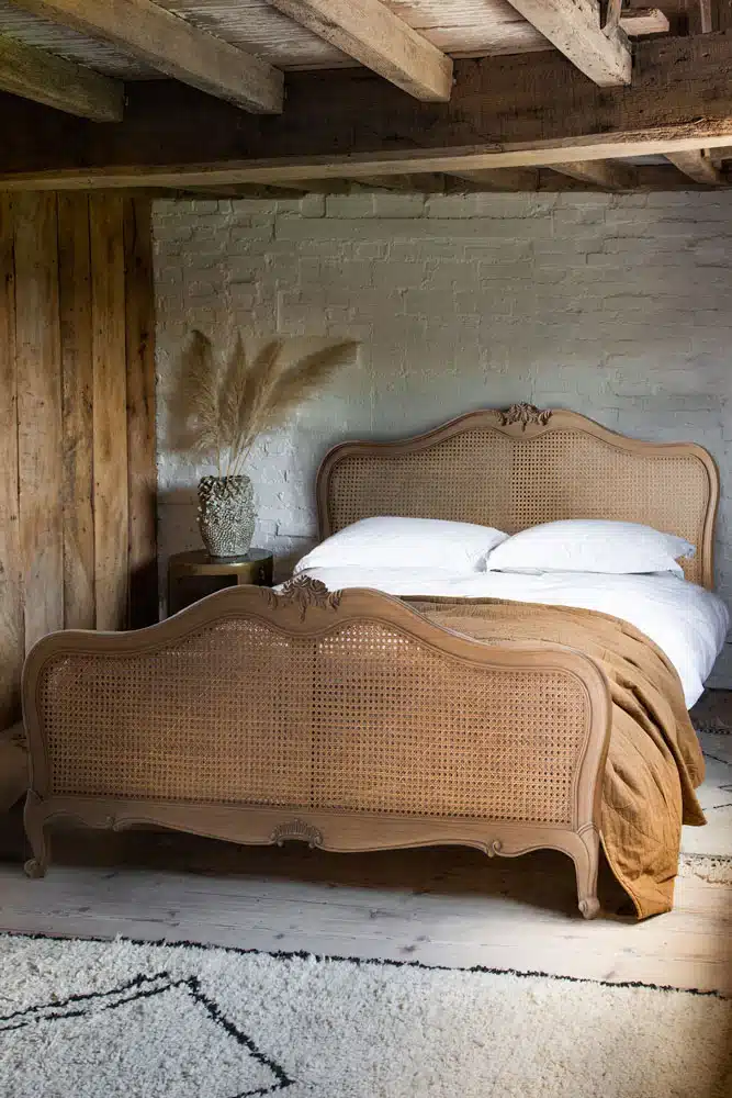 Rustic beige bedroom with a French-style cane bed, exposed timber ceiling beams, whitewashed brick wall, ochre linen throw and a Berber rug