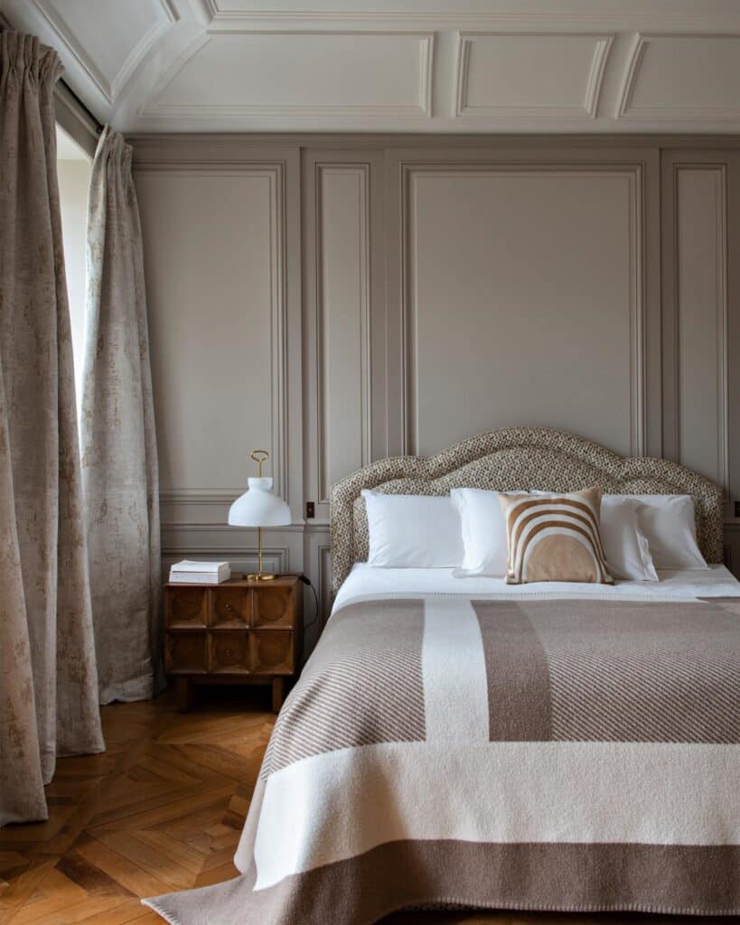 Luxury beige bedroom with Farrow & Ball Skimming Stone boiserie panelling, herringbone parquet floor, upholstered headboard, carved walnut bedside table and striped wool throw. Image: @juliette_saier via Farrow & Ball
