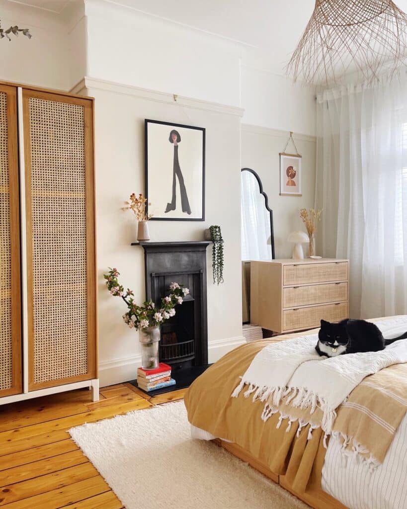 Farrow & Ball Slipper Satin beige bedroom with cane wardrobe, rattan chest of drawers, black Victorian fireplace, arch mirror, mustard throw and pine floorboards. Image: @1930s_semi_charmed_life via Farrow & Ball