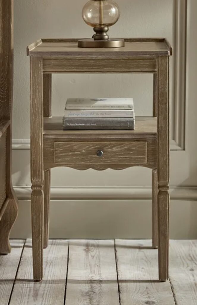 Oak-toned Louves bedside table with open shelf and slim drawer, styled with beige bedroom decor for a rustic yet refined look.