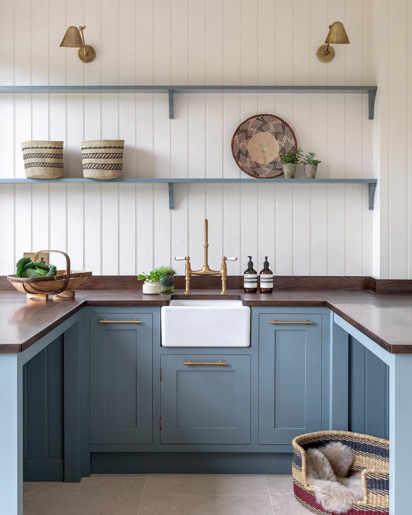 De Nimes soft blue-grey lower kitchen cabinets with dark wood worktop, white Belfast sink, brass bridge tap, open shelving and tongue and groove wall panelling.