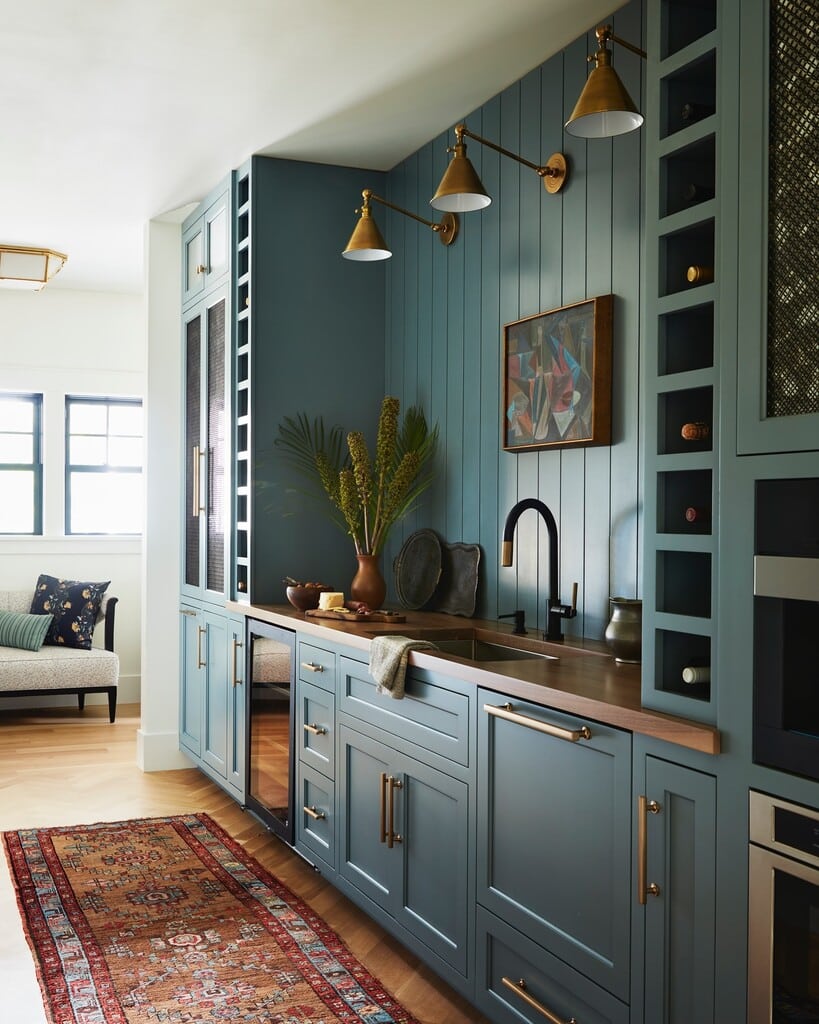 De Nimes full-height kitchen cabinets with integrated open shelving, wine storage, dark wood worktop, brass bar handles, brass wall lights and Persian rug.​​​​​​​​​​​​​​​​