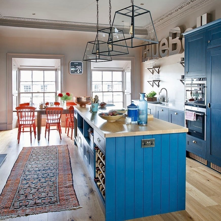 Hague Blue open-plan kitchen with Charleston Gray walls, butcher block island top, coral red dining chairs, Persian rug and industrial cage pendant light.