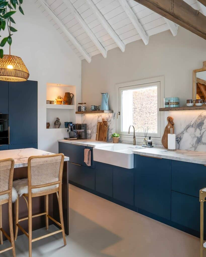 Hague Blue handleless kitchen cabinets with marble worktop and backsplash, vaulted white ceiling with exposed beams, rattan bar stools and Belfast sink.