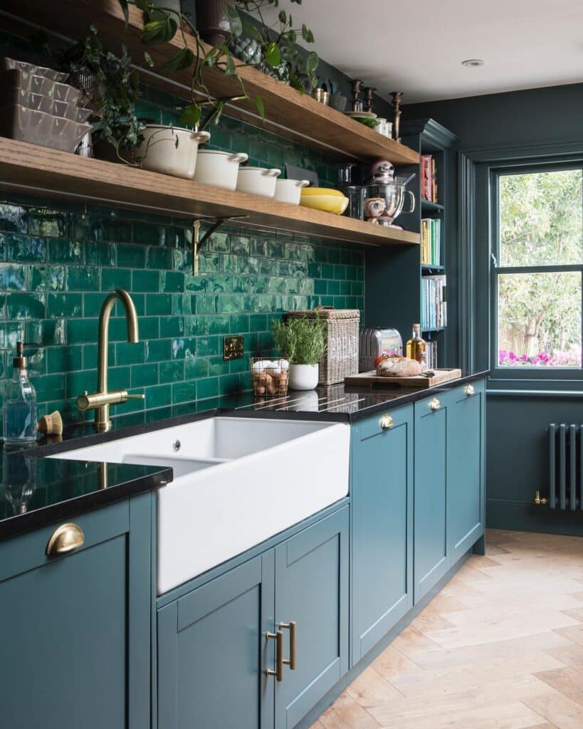 Inchyra Blue shaker kitchen cabinets with emerald green glazed brick tile backsplash, white Belfast sink, brass tap and open oak shelving with plants.